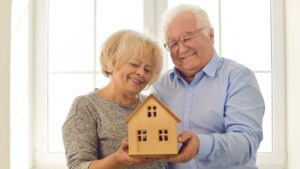 A happy couple holding a wooden house, celebrating paying off their mortgage and achieving financial freedom. Ballentine Capital Advisors, a trusted wealth management firm in Greenville, SC, helps clients pay down debt, build emergency funds, and secure their financial future.