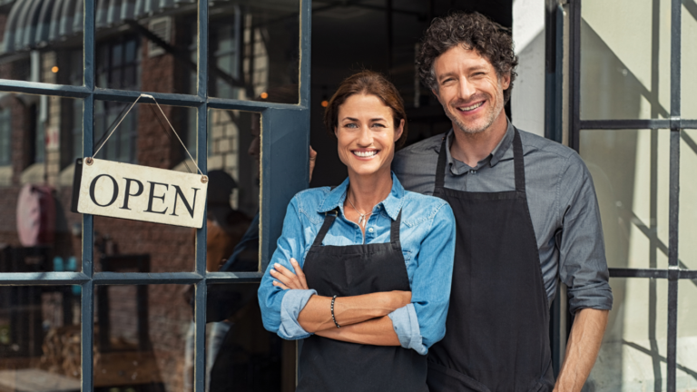 smiling couple standing in front of an "Open" sign, representing small business ownership and financial planning for 2025. Ballentine Capital Advisors, a wealth management firm in Greenville, SC, helps small business owners navigate tax changes under the new administration, maximize IRS contributions, and plan for long-term financial success.